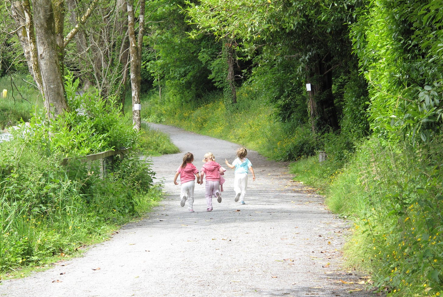 Three young children run on a path in a wooded area
