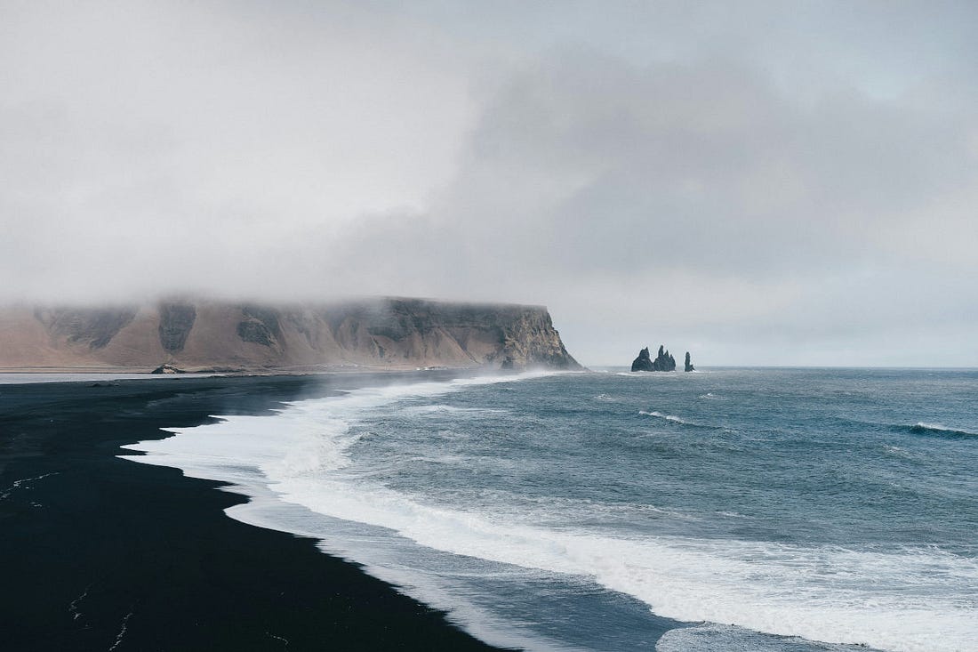 una playa negra de Islandia, batida por el mar, con un acantilado al fondo que se pierde en las nubes una playa negra de Islandia, batida por el mar, con un acantilado al fondo que se pierde en las nubes