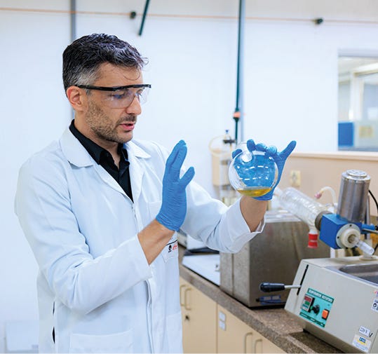 Um homem de cabelos curtos e grisalhos veste jaleco branco, camisa preta, óculos de proteção e luvas azuis. Ele está em um laboratório, segurando um balão de vidro com um líquido amarelado e gesticulando com a outra mão. Ao fundo, veem-se bancadas e equipamentos científicos, como um evaporador rotativo. A imagem é bem iluminada e foca na ação do pesquisador.