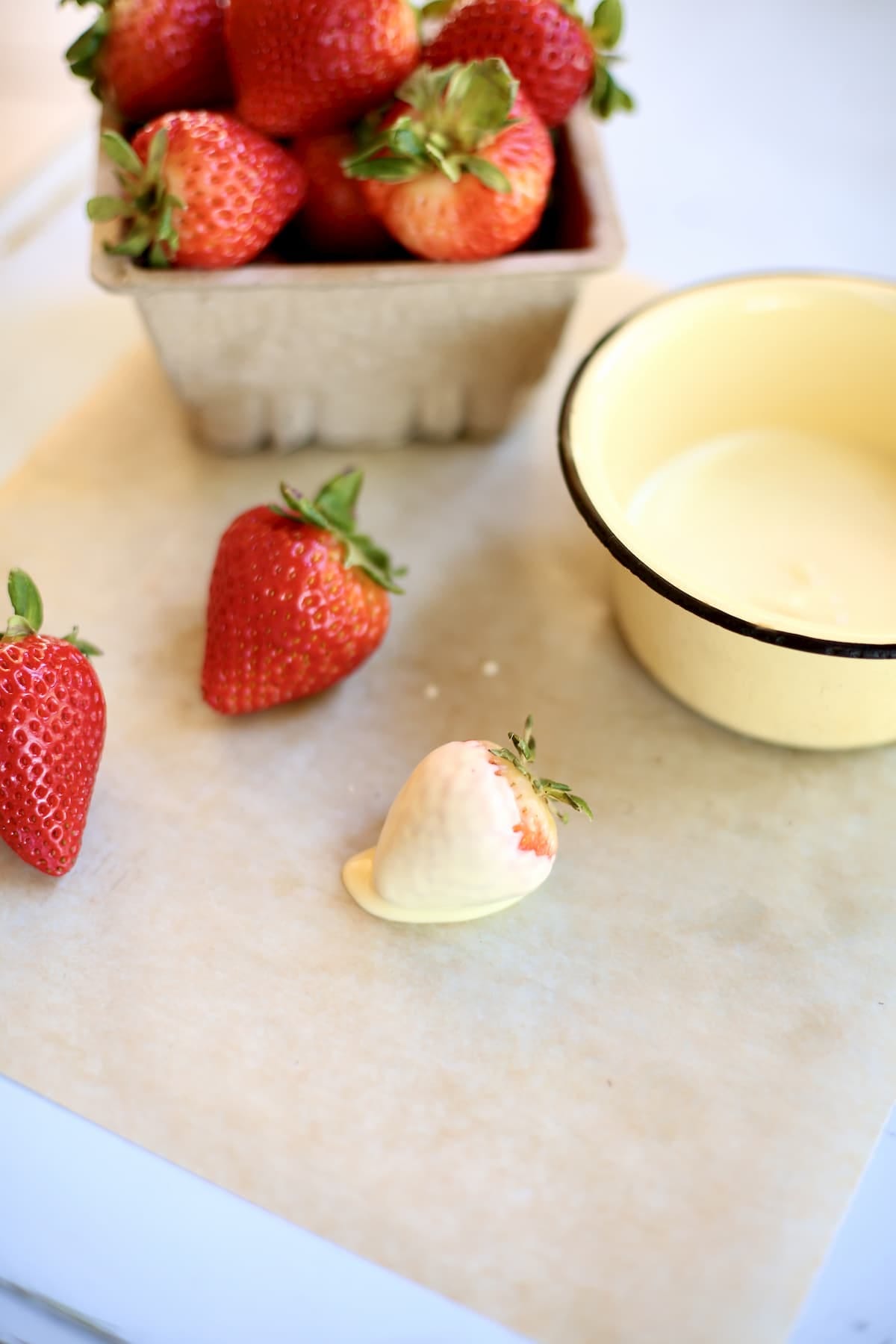 a set up of white chocolate and strawberries, one after being dunked and parchment paper a set up of white chocolate and strawberries, one after being dunked and parchment paper