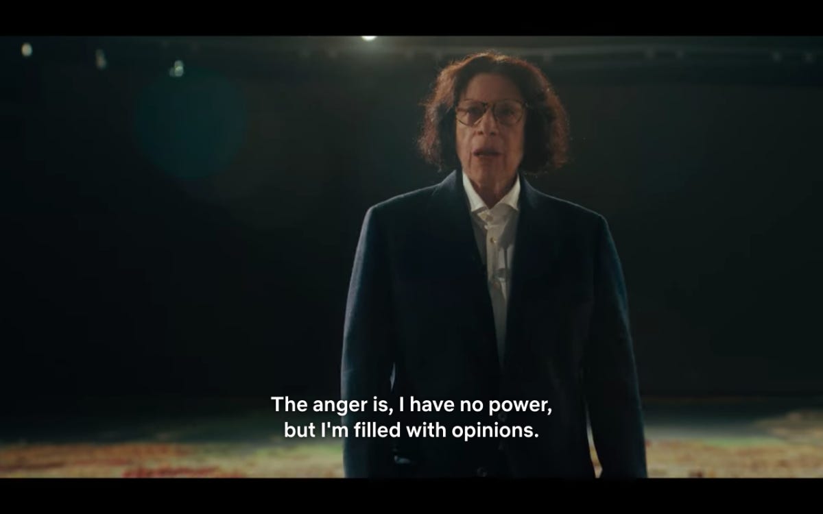 A film still of Fran Lebowitz standing slightly off center in front of a miniature replica of the city of NY. She has wearing her signature white button down and blazer, with glasses. In white text on the bottom of the screen it says "The anger is, I have no power, but I'm filled with opinions."
