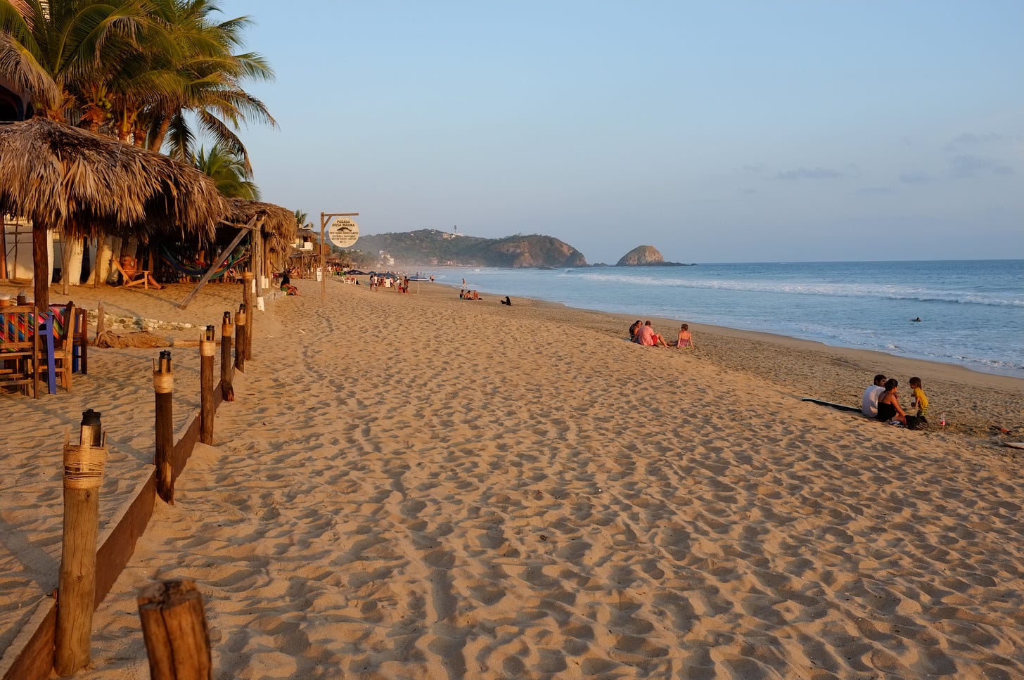 A wide view of Zipolite Beach at sunset, with gentle waves, golden sand, scattered groups of people sitting near the shoreline, and palapa-covered beach bars and hammocks lining the left side of the frame. A wide view of Zipolite Beach at sunset, with gentle waves, golden sand, scattered groups of people sitting near the shoreline, and palapa-covered beach bars and hammocks lining the left side of the frame.