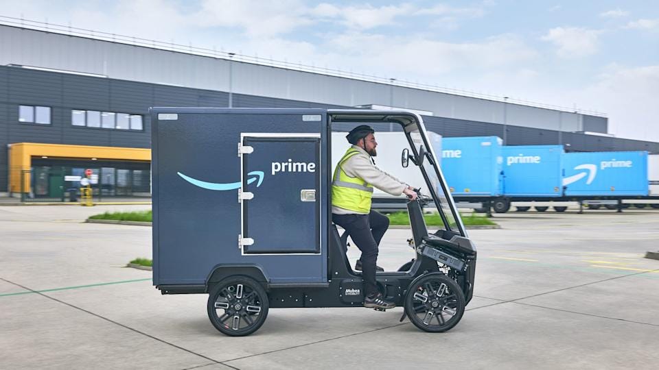 A driver operates a dark-blue Amazon e-cargo bike with an Amazon warehouse and light-blue trucks in the background.