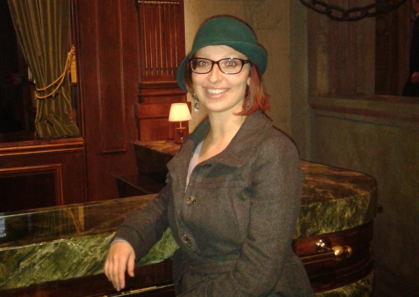 Elena che guarda in camera, sorridendo. Ha un cappello asimmetrico verde, occhiali rettangolari, un cappotto grigio. Si appoggia a un bancone di marmo. Elena che guarda in camera, sorridendo. Ha un cappello asimmetrico verde, occhiali rettangolari, un cappotto grigio. Si appoggia a un bancone di marmo.