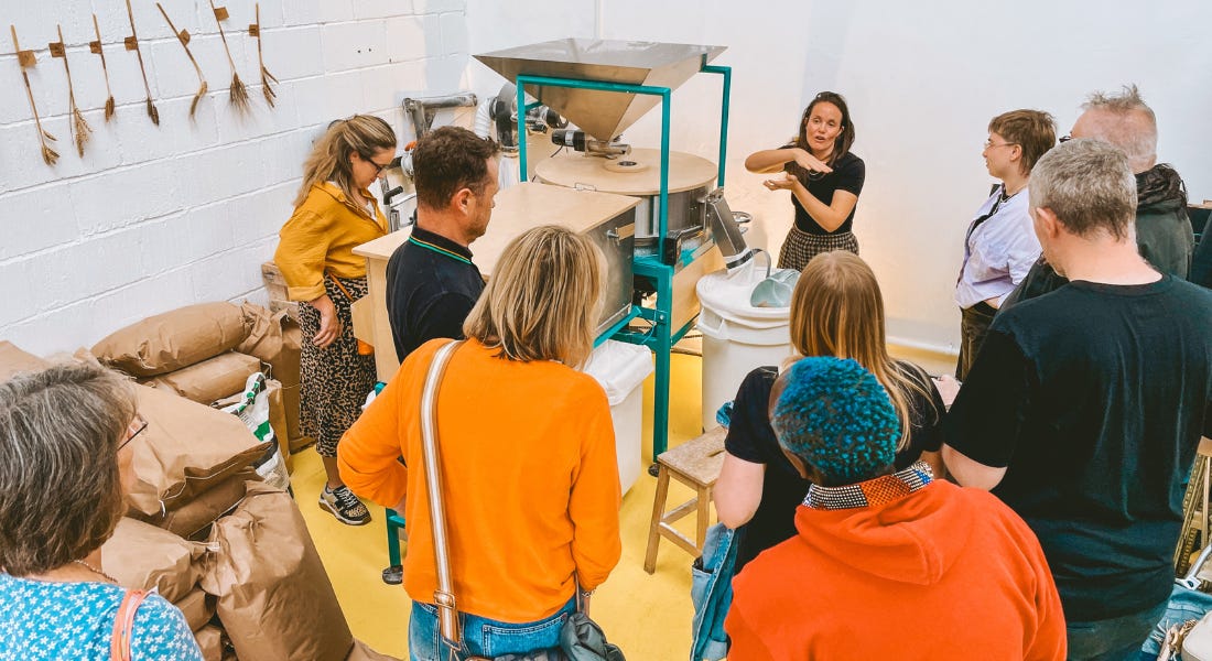 A group of people standing around a flour milling machine listening to a woman speak