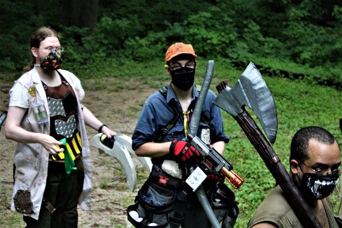 4 cowled and masked figures at night holding a finger over their mouth shushing the camera. A LARPer striking someone dressed as a zombie, exposed ribs and gorey makeup, with a foam sword. LARPers standing in the woods. One wears a torn lab coat and reinforced leather armor, and another in an orange baseball cap weilds a nerf gun and has too many tool belts. That's me, with the orange baseball cap. A third is mostly out of frame, but their large foam axe sticks up into view. A collection of LARPers stand outside a cabin with "RH" on the wall and a pop-up tent outside. Weapons and gear lay around the area and on the table.