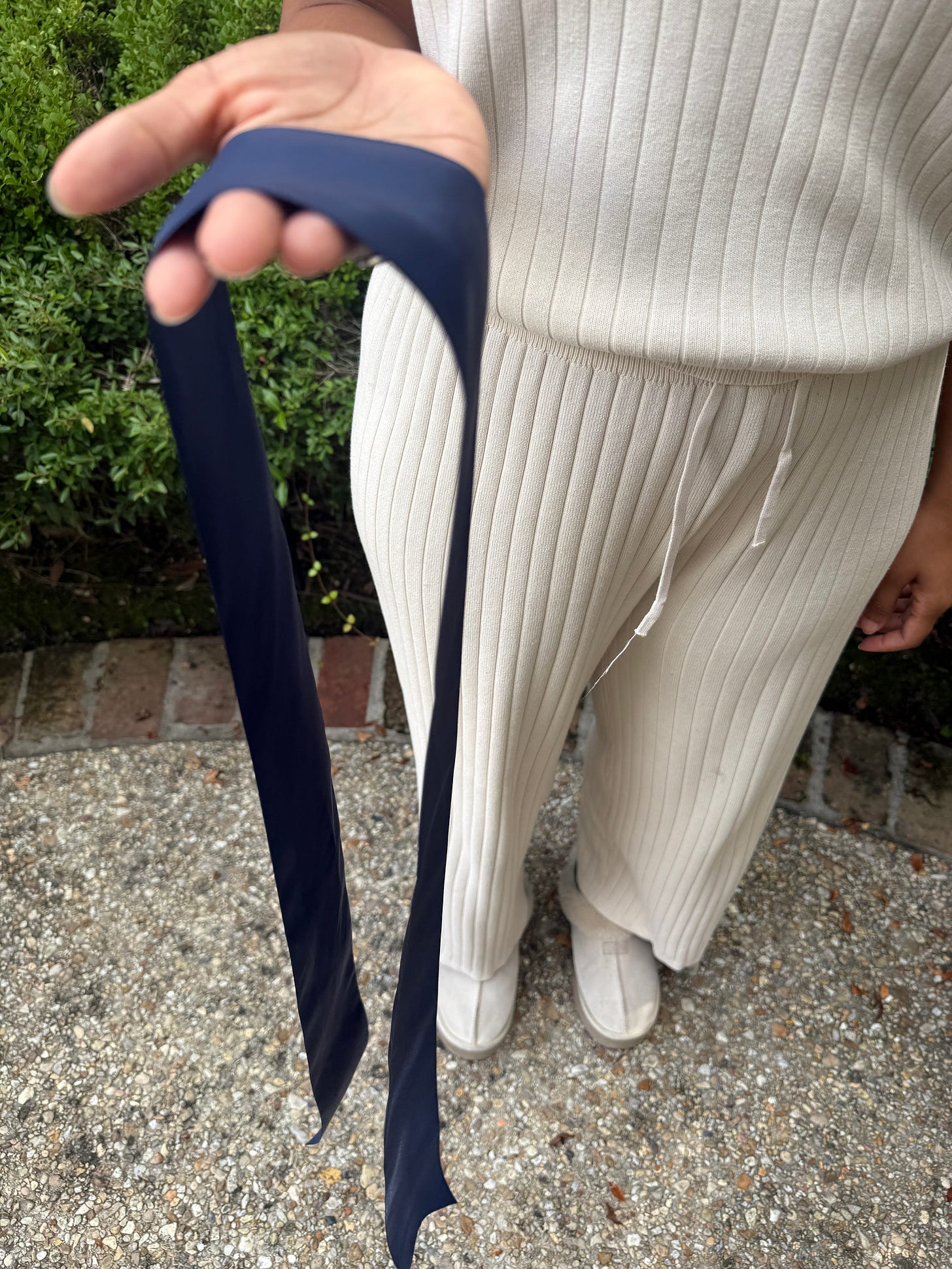 Hands holding a navy blue satin ribbon against white ribbed pants, demonstrating the first step of bow tying