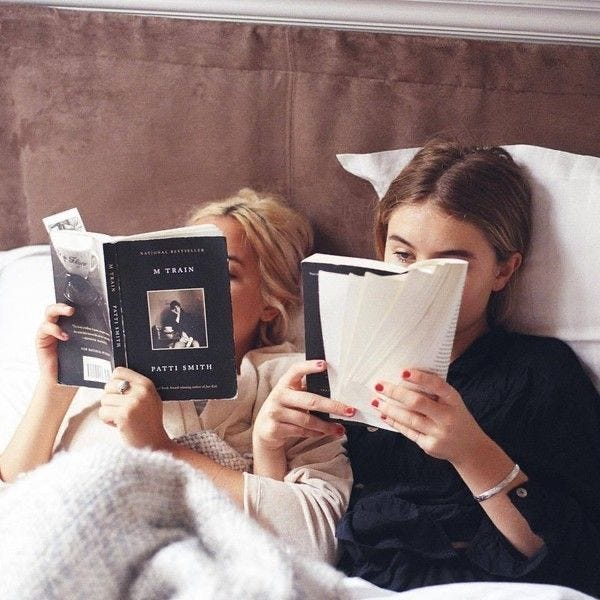 This may contain: two women laying in bed reading books and smiling at the same person's face