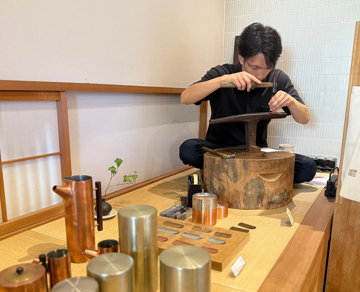 A person sits on a platform, using a hammer and metal tool to work on a dark metal object. Various metal items and tools are displayed on the table in front.
