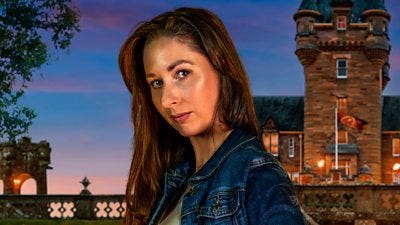 A woman with long brown hair looks to camera, side on, wearing a denim jacket and green top. She's set against the backdrop of a castle by night. A woman with long brown hair looks to camera, side on, wearing a denim jacket and green top. She's set against the backdrop of a castle by night.