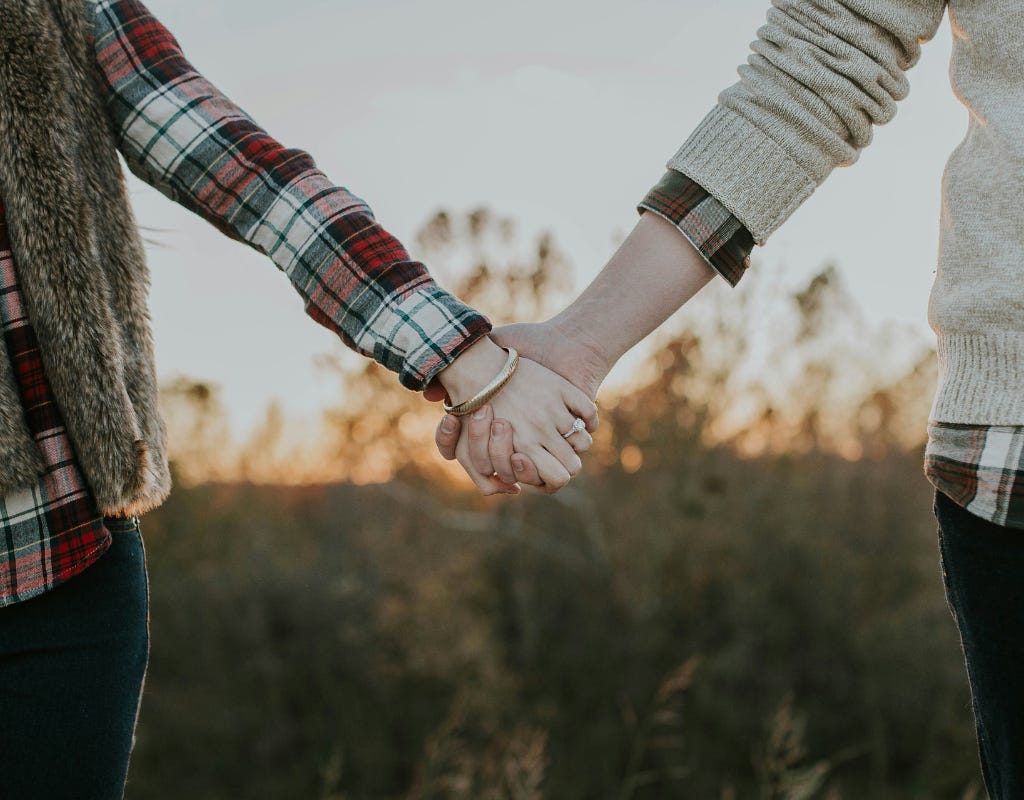 Two persons, each wearing blue jeans and a red, white and green plaid flannel shirt, are  holding hands.  One in the couple is wearing a short fur vest over the flannel shirt, and has a white diamond ring on the ring finger, as well as a gold bracelet on the same hand.  The other person is wearing a beige sweater over the flannel shirt, with the cuffs of the sweater pulled up to reveal a small part of the flannel shirt.  There is a field of out-of-focus tall green grass below their clasped hands between them.