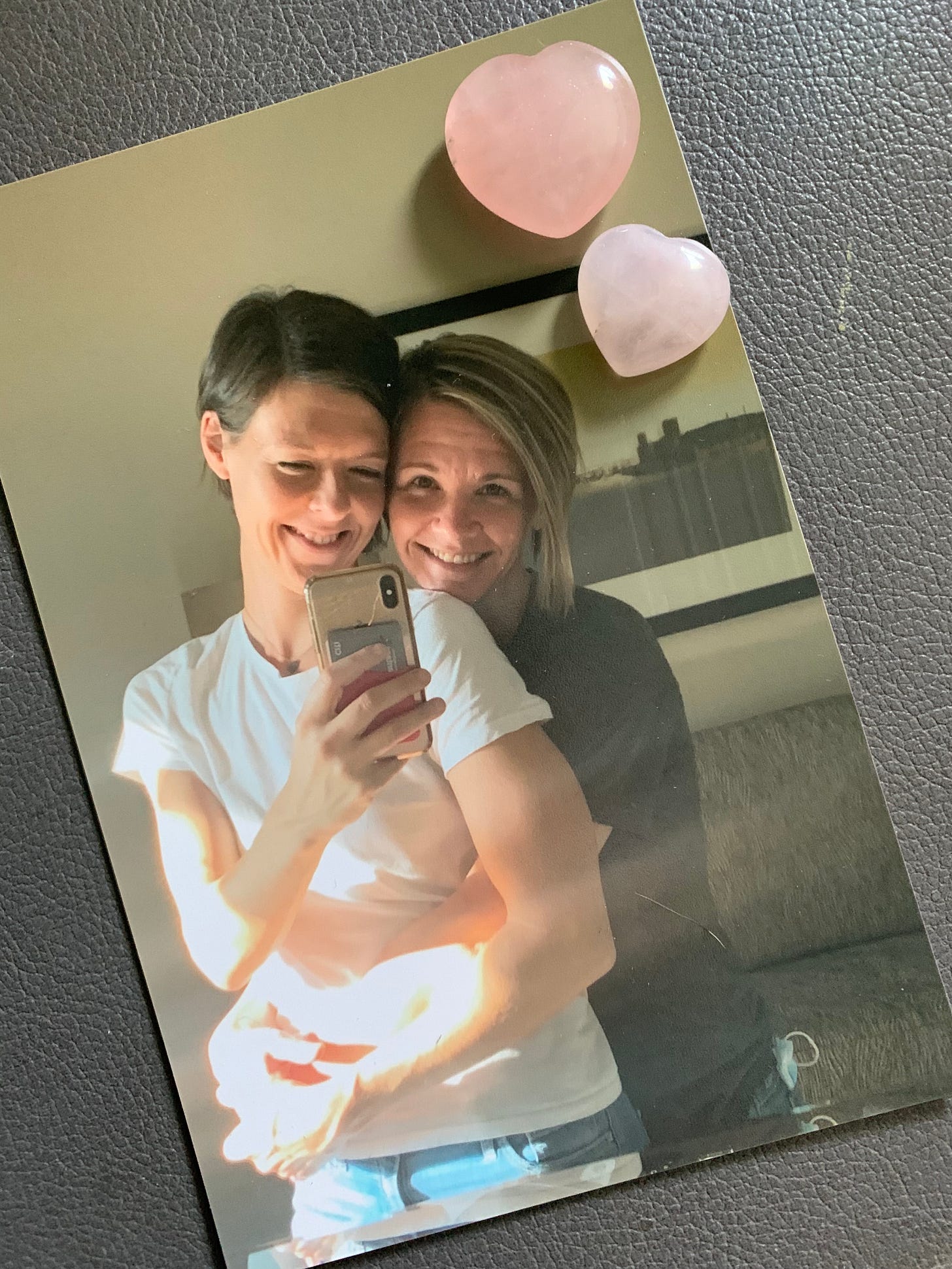 Photo graph of two women with short hair smiling and embracing. Two pink stone hearts sit at the top right corner of the photo.