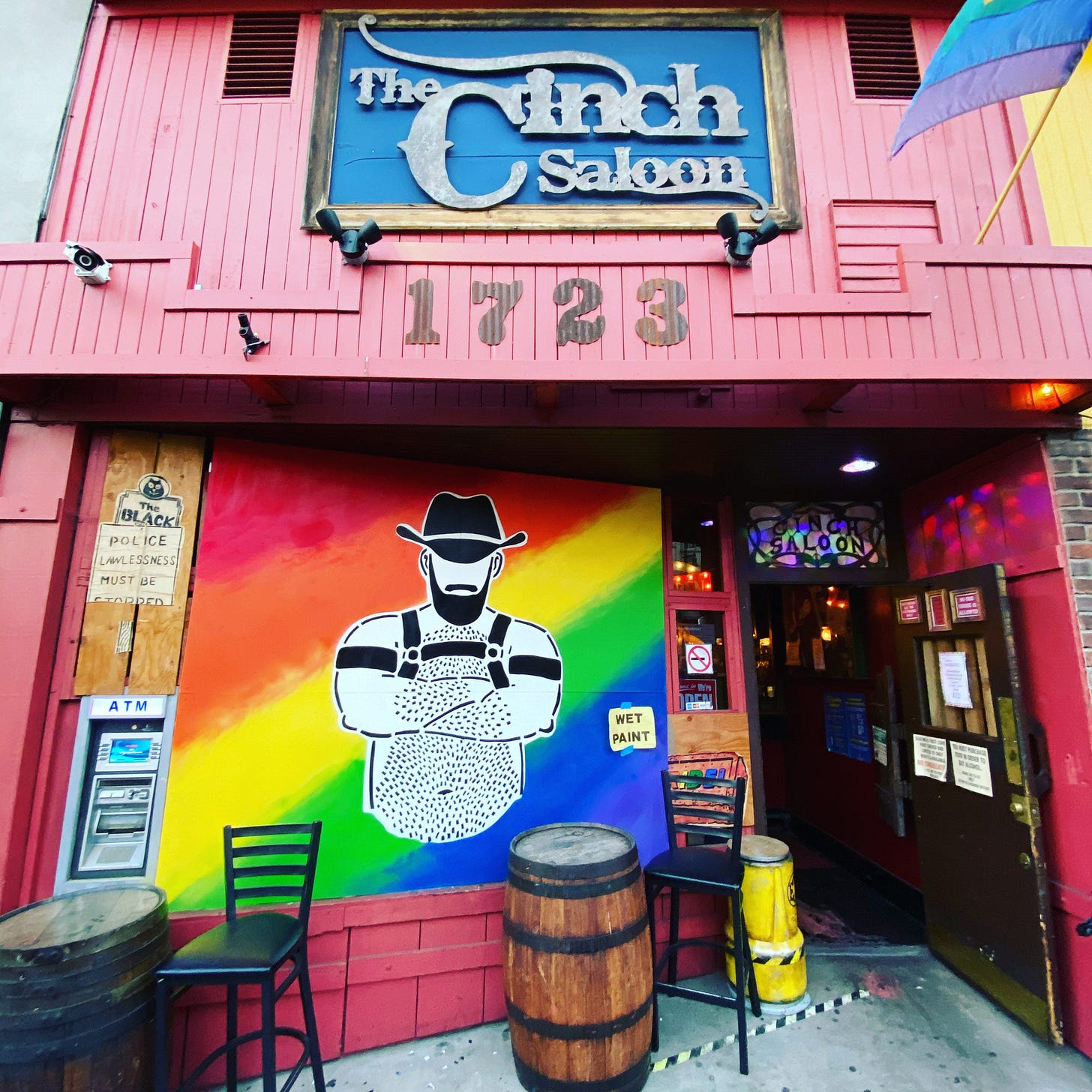 Front of the Cinch Saloon with a colorful mural of a gay cowboy on a rainbow background. Bright, lively atmosphere with chairs and barrels outside. The Cinch Saloon has a blue sign, with a pink building, and the number 1723 on top. Jeremy's work of the cowboy is in the front. He is shirtless, wearing a chest harness and black arm bands. He has a hair chest and arms, and a black beard and black cowboy hat. There is another work of Jeremy's to the left, of the Black Nite brawl. There is an ATM on the left side, by one of the barrel tables. The Cinch Saloon's door is open, and there is a rainbow flag flying over it. There is also a wet paint sign on the mural. Front of the Cinch Saloon with a colorful mural of a gay cowboy on a rainbow background. Bright, lively atmosphere with chairs and barrels outside. The Cinch Saloon has a blue sign, with a pink building, and the number 1723 on top. Jeremy's work of the cowboy is in the front. He is shirtless, wearing a chest harness and black arm bands. He has a hair chest and arms, and a black beard and black cowboy hat. There is another work of Jeremy's to the left, of the Black Nite brawl. There is an ATM on the left side, by one of the barrel tables. The Cinch Saloon's door is open, and there is a rainbow flag flying over it. There is also a wet paint sign on the mural.