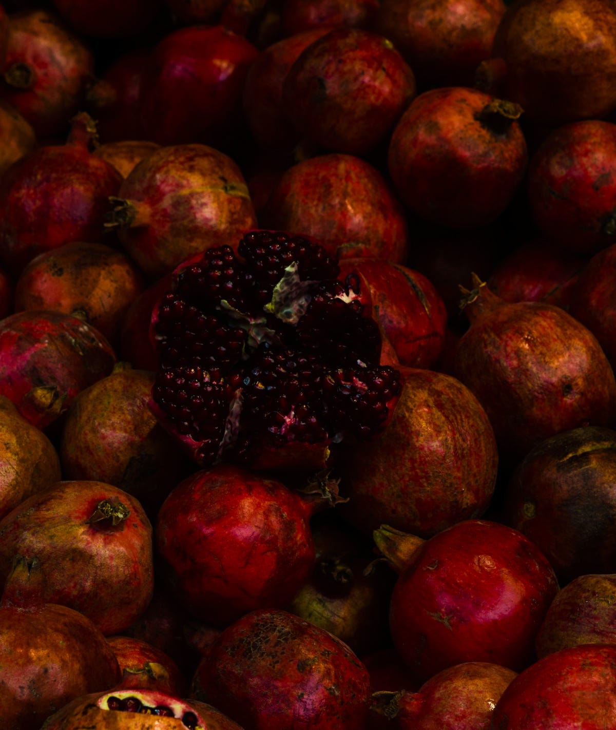 Dark, high contrast, somber perspective from above on a pile of pomegranates, one is split open, displying its seedlings messily.
