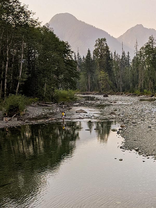 man in yellow shirt carries a child across a placid shin-deep river below a mountain vista tinted pink by smoke