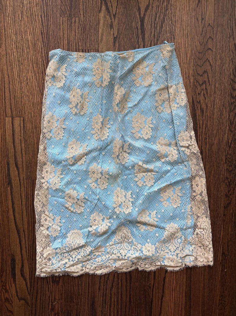 May include: A light blue skirt with a floral lace overlay. The skirt features a delicate, cream-colored lace pattern and a scalloped hemline. The skirt is made of a lightweight material.