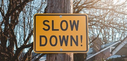Yellow sign on wooden post that reads Slow Down! In the background there are leafless trees, house roofs, and sunshine.