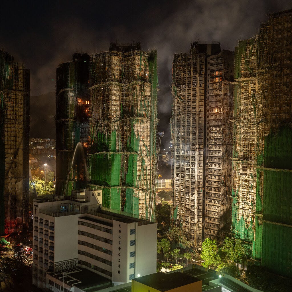 High-rise buildings, many wrapped in green scaffold netting, are illuminated at night. A fire rages in one, with a stream of water visible.
