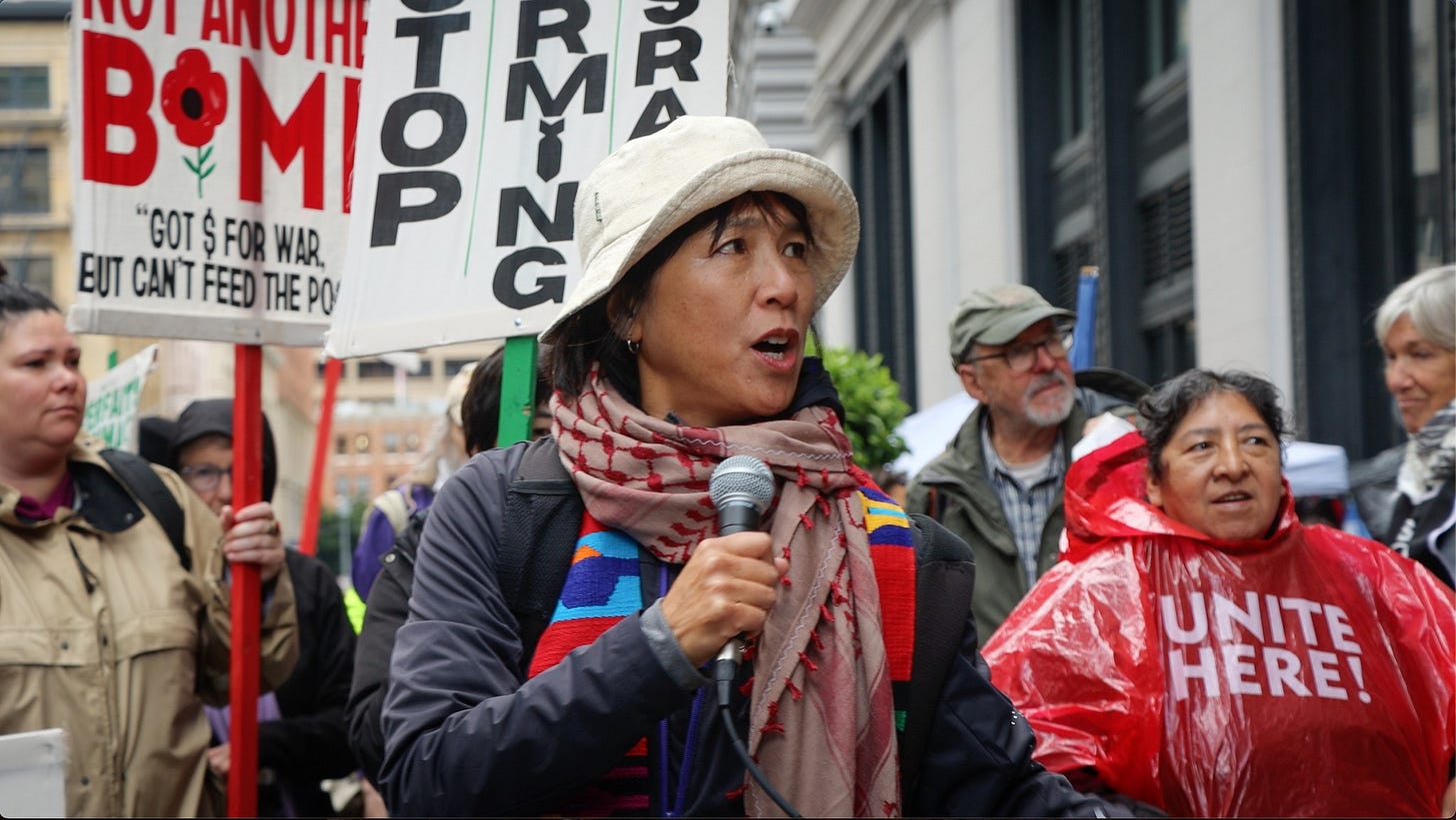 Rev Deborah at a march for Palestinian liberation on a city street. People carry signs that read: "Not Another Bomb," "Stop Arming Israel," Unite Here," and are dressed in rain gear.