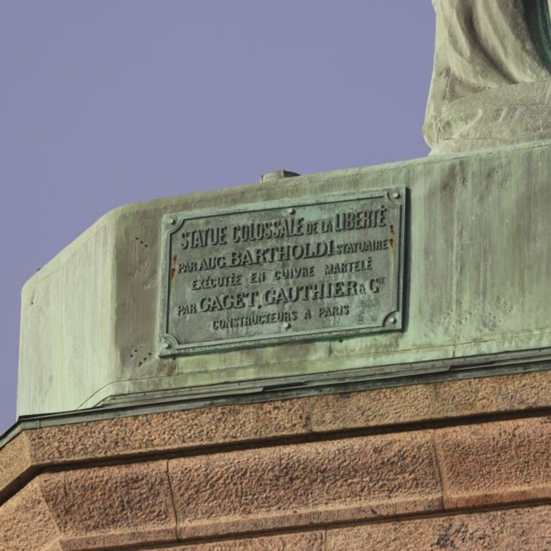 Green copper plaque with French words. The plaque says: "Statue colossale de la liberté par Aug. Bartholdi  Statuaire. Exécutée en cuivre martelé par Gaget, Cauthier, &  Cie. Constructeurs a Paris."

Translated into English, the plaque says:

"Colossal Statue of Liberty by Aug. Bartholdi, statue maker. Made in hammered copper by Gaget, Cauthier and Company, builders in Paris"