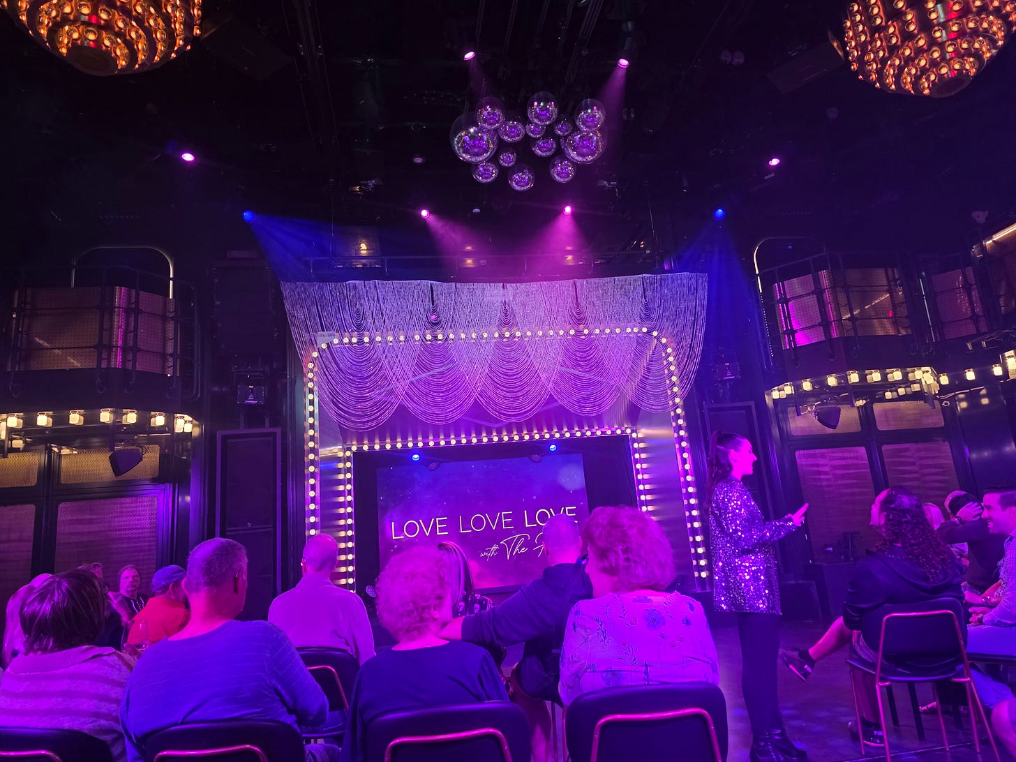 A look inside The Manor, ten or so rows of people in loose seats looking toward a stage that has a sign that says, "Love, Love, Love". The lighting is purple and atmospheric