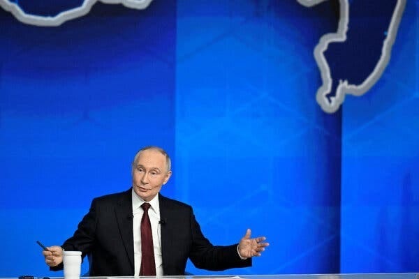 President Vladimir Putin of Russia, in a black suit and a dark red tie, speaking and gesturing in front of a blue backdrop.