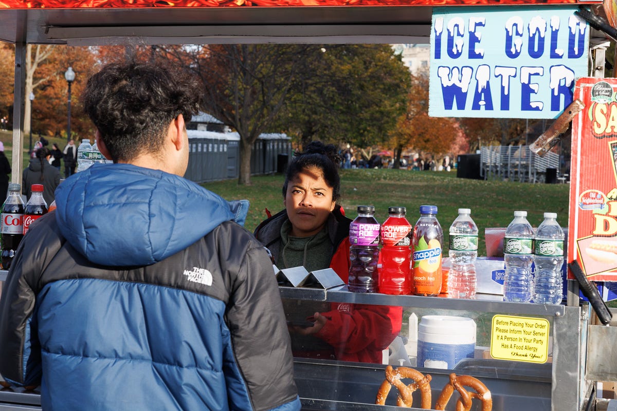 vendor behind a food cart listening to a customer as sunlight hits rows of drinks