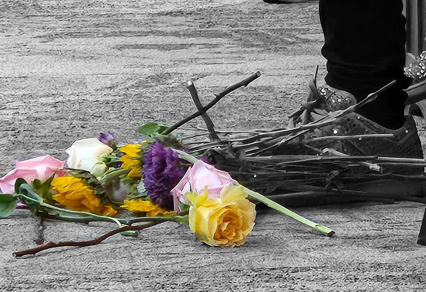 A bunch of cut flowers lie on the floor by a person's sneaker-clad foot. The flowers are in color while the rest of the photo is black and white.
