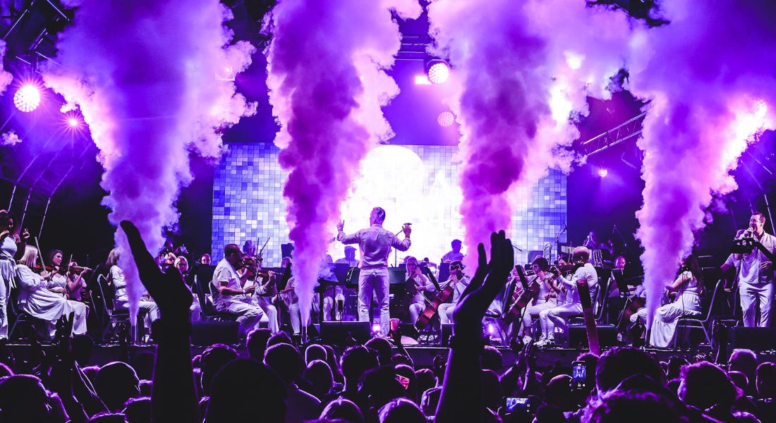 A concert with a stage with a live orchestra, a large crowd, puffs of smoke, and purple lights