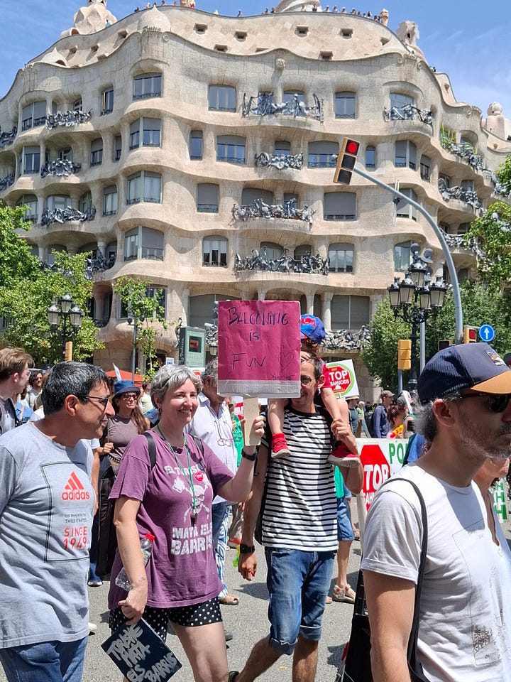 1.Una donna sorridente con capelli grigi corti e una maglietta viola tiene in mano un piccolo cartello rosa con la scritta "BALCONING IS FUN" (Il balconing è divertente) barrata da una riga. Di fianco, un uomo porta un bambino a spalle. Dietro di loro, si erge la facciata ondulata e iconica della Casa Milà (La Pedrera) di Gaudí. La scena si svolge all'aperto, durante una manifestazione 2.Due persone in primo piano tengono dei cartelli durante una manifestazione. La persona a sinistra, che indossa una maglietta grigia e pantaloncini chiari, tiene un grande cartello orizzontale in cartone. Sul cartello, c'è scritto "HABITANTS 1.62 MILIONS" (Abitanti 1.62 milioni) e "TURISTES 26.1 MILIONS" (Turisti 26.1 milioni), con un segnale di "STOP" rosso tra le due cifre. Sotto, in lettere più grandi, si legge "TURISME MASSIU" (Turismo di massa). Accanto a questa persona, c'è un cane chiaro al guinzaglio. La persona a destra, che indossa una canottiera scura, pantaloni chiari e una borsa a tracolla, tiene un cartello verticale sempre in cartone con la scritta "TOURISM IS KILLING BARCELONA" (Il turismo sta uccidendo Barcellona) sopra un'immagine stilizzata di fiamme e la frase "DECREIXEMENT TURISTIC JA!!" (Decrescita turistica ora!!) in basso. Sullo sfondo si intravedono altre persone che partecipano alla manifestazione e alberi lungo la strada.