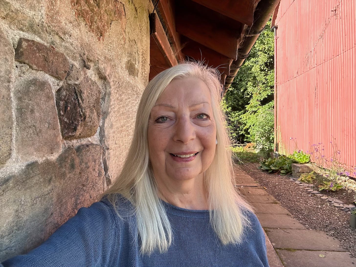 Head and shoulders images of Yasmin Chopin a mature woman with long grey hair stands beside a stone building in the Scottish Borders of the UK