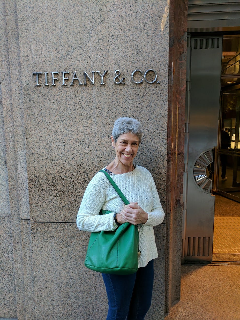 A white woman with white hair holds a green purse in front of a Tiffany sign A white woman with white hair holds a green purse in front of a Tiffany sign