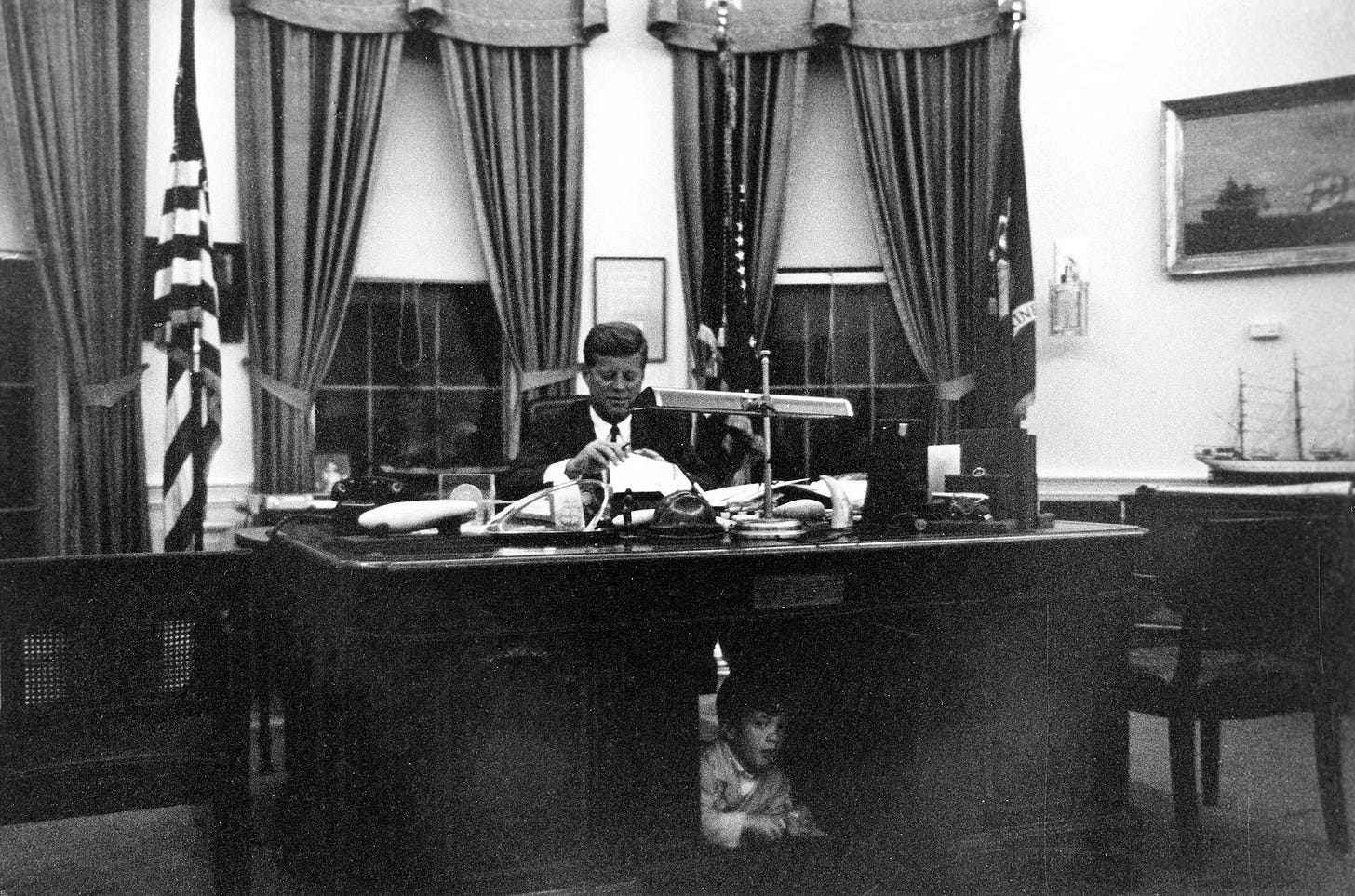 File handout image shows former U.S. President Kennedy at his White House Oval Office desk while son JFK Jr. looks out from underneath