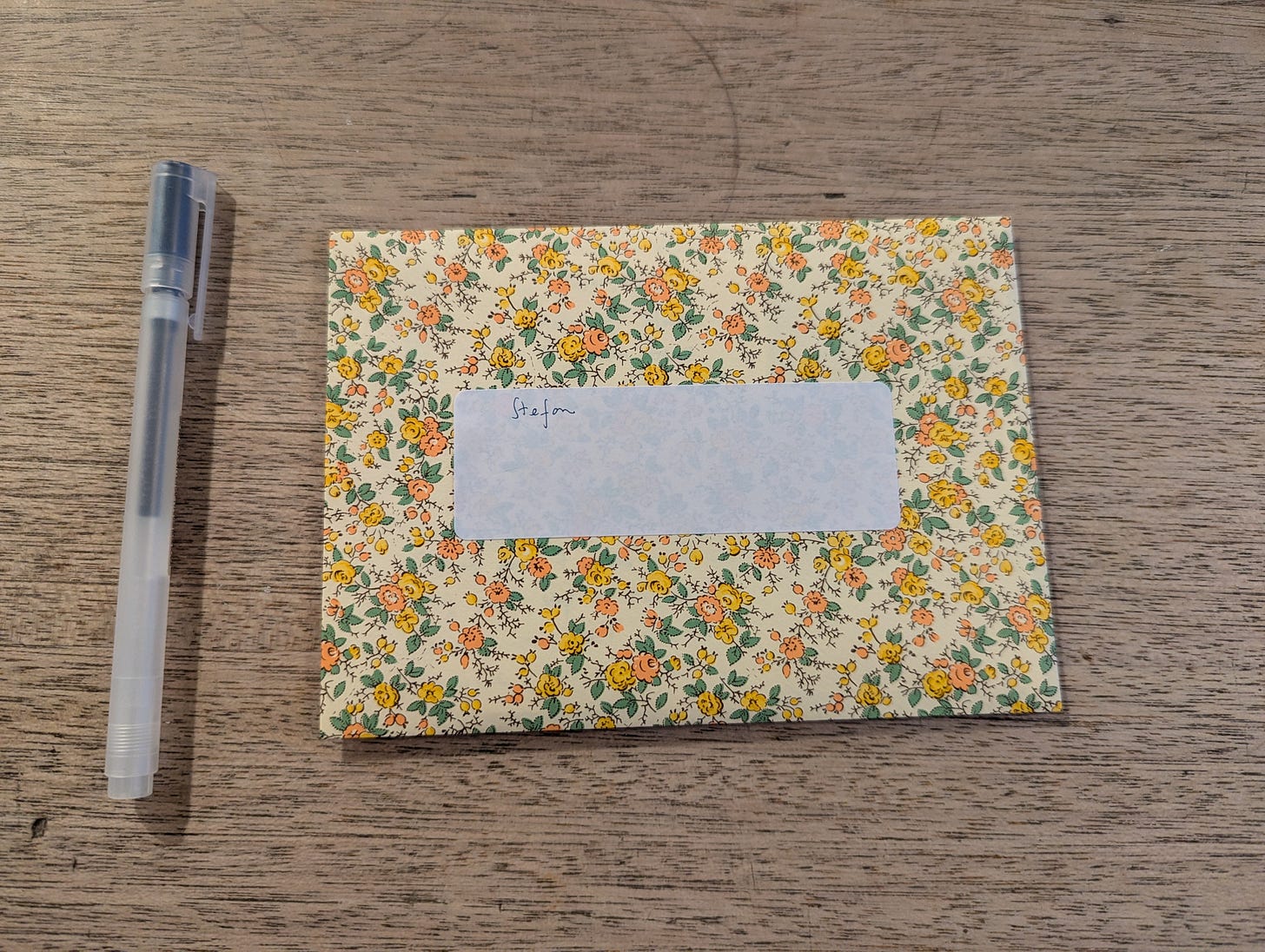 An envelope with yellow and orange floral pattern on cream background, addressed to 'Stefan' in handwritten script. A grey pen lies beside it on a wooden table surface.