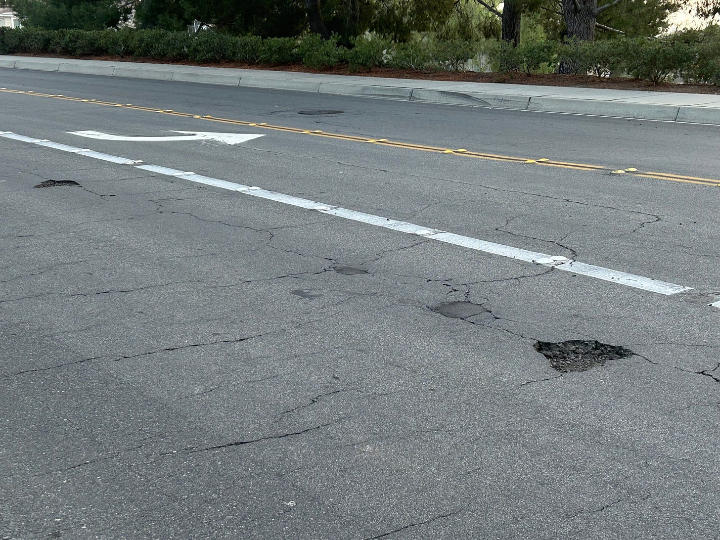 A wide asphalt road webbed with cracks and pitted with potholes.