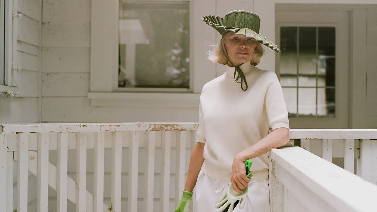 Pamela Anderson in white outfit green hat holding spade basket tote leaning on white fence of her garden