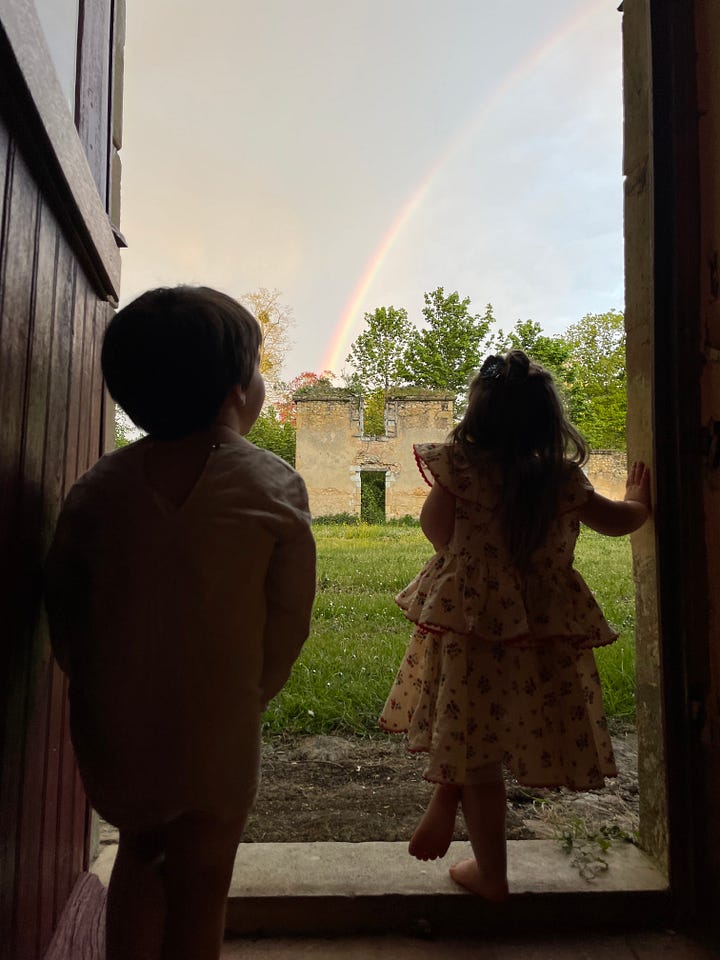 A collage of the grounds around the rented farmhouse, including an iron gate set into an ivy-covered stone wall, a garden path, a courtyard strung with bunting at dusk, two small children watching a rainbow from a doorway, pink cyclamen carpeting the ground beneath a large tree, and children walking down tree-lined country paths.