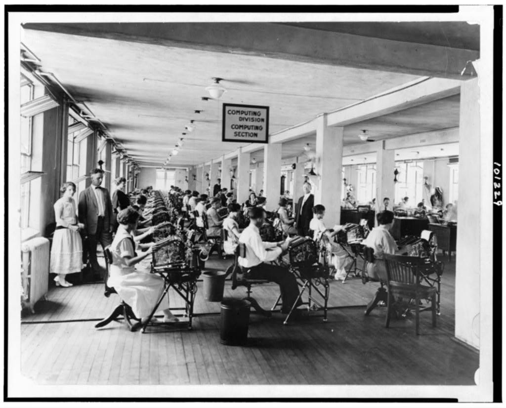 Archival black and white image of men and women at calculating desks, early 20th centruy, with a sign saying "Computing Division" Archival black and white image of men and women at calculating desks, early 20th centruy, with a sign saying "Computing Division"