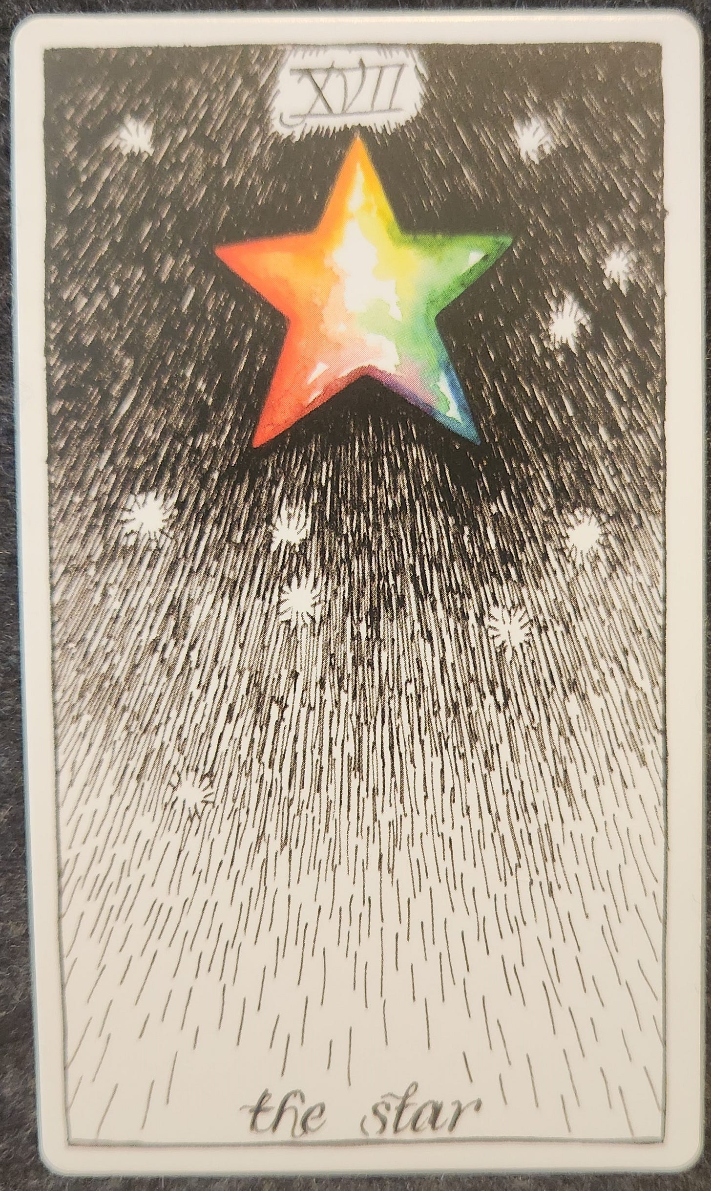 A tarot card labelled XVII the star the tower. A rainbow coloured star sits in the middle of a dark night sky with a few scattered white stars surrounding it. 