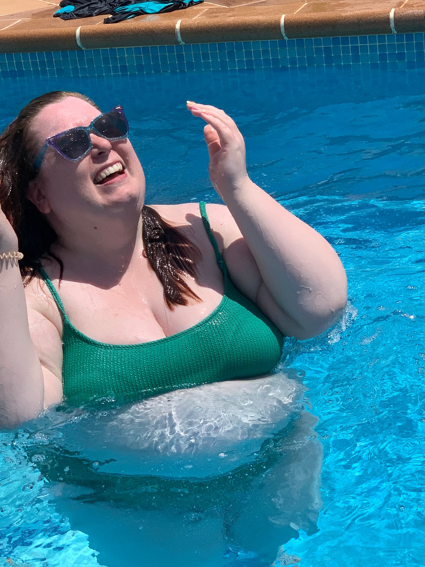Photo of Melanie, a plus size woman, wearing sunglasses and a green bikini, standing in a pool smiling for the camera