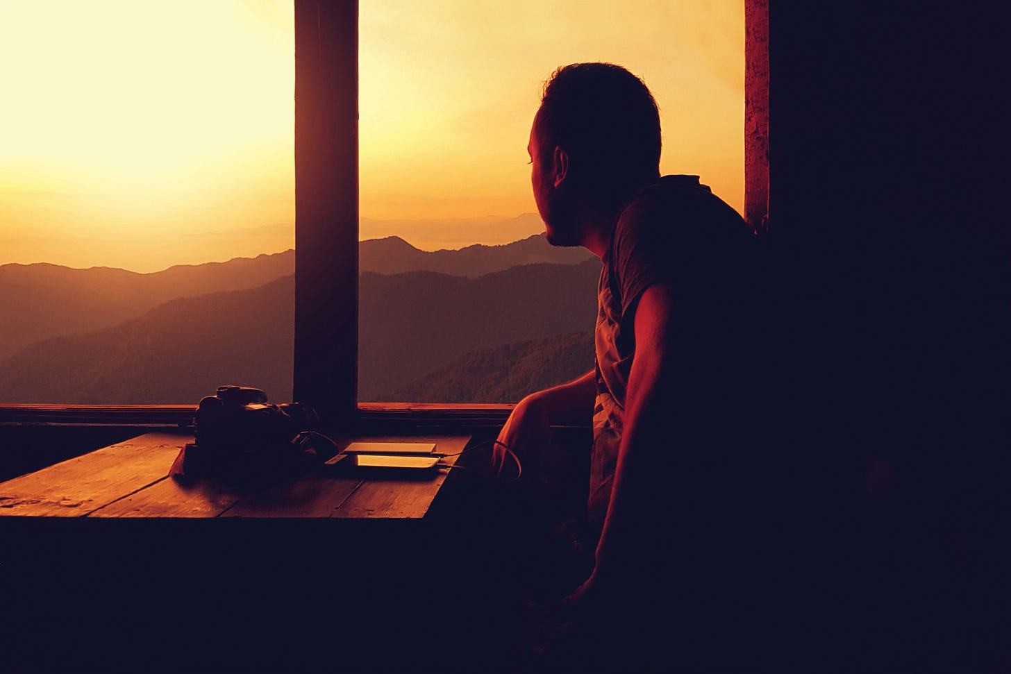 A man sits in silhouette at a wooden desk, looking out through a large window toward a mountain range bathed in golden sunset light.