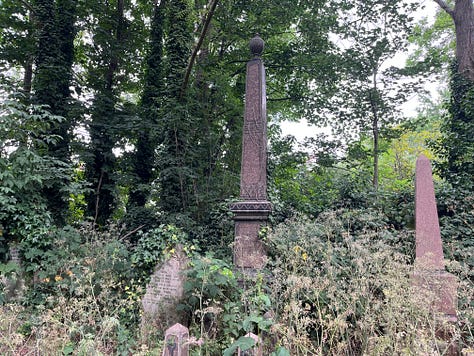 Three obelisk grave monuments with adornments. One has a hand pointing upwards on top, one a laurel wreath carved into the front, and one has an urn on top.