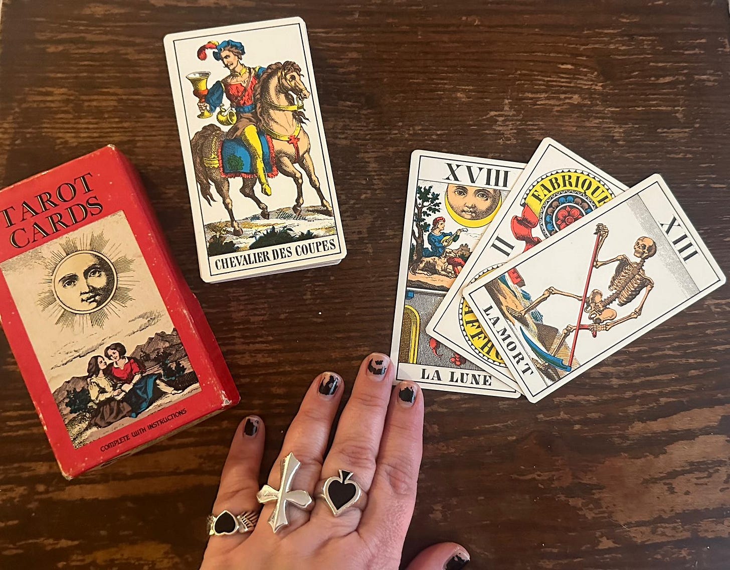 Tarot de Marseille cards on wooden table including Chevalier des Coupes (Knight of Cups), La Lune (The Moon XVIII), La Mort (Death XIII) and La Roue de Fortune (Wheel of Fortune X), beside vintage tarot card box and hand with rings, photographed for the Tarot DMs interview with Alice Slater by Finbarre Snarey.