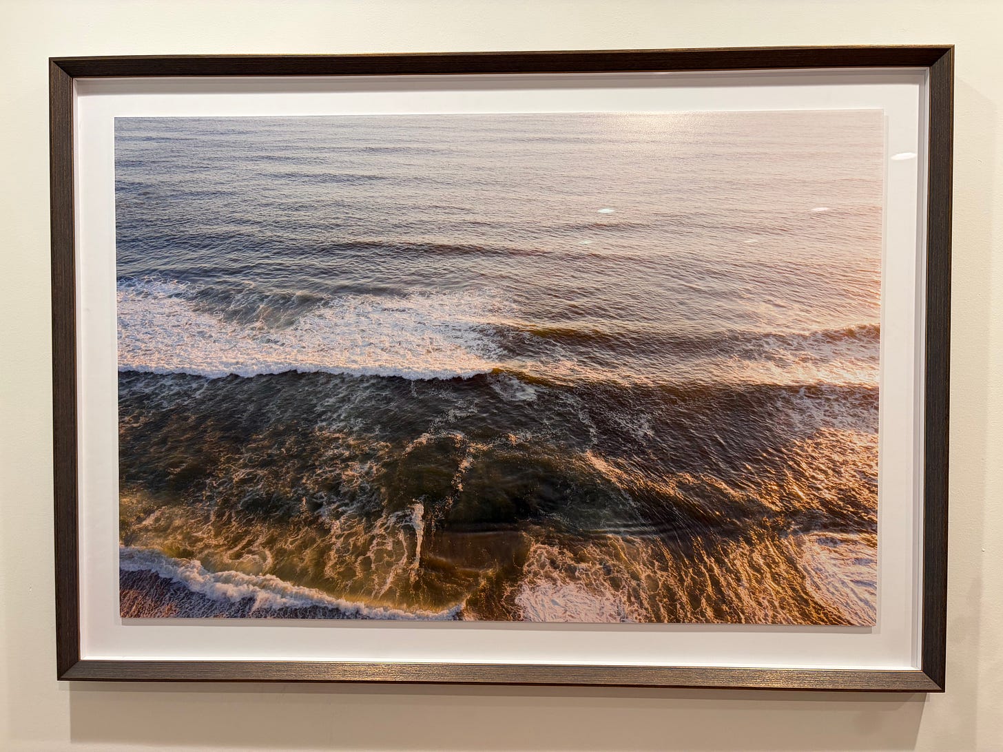 Close view of an ocean photograph showing waves, foam, shadow, and warm light inside a dark walnut frame with white space around the print. Close view of an ocean photograph showing waves, foam, shadow, and warm light inside a dark walnut frame with white space around the print.
