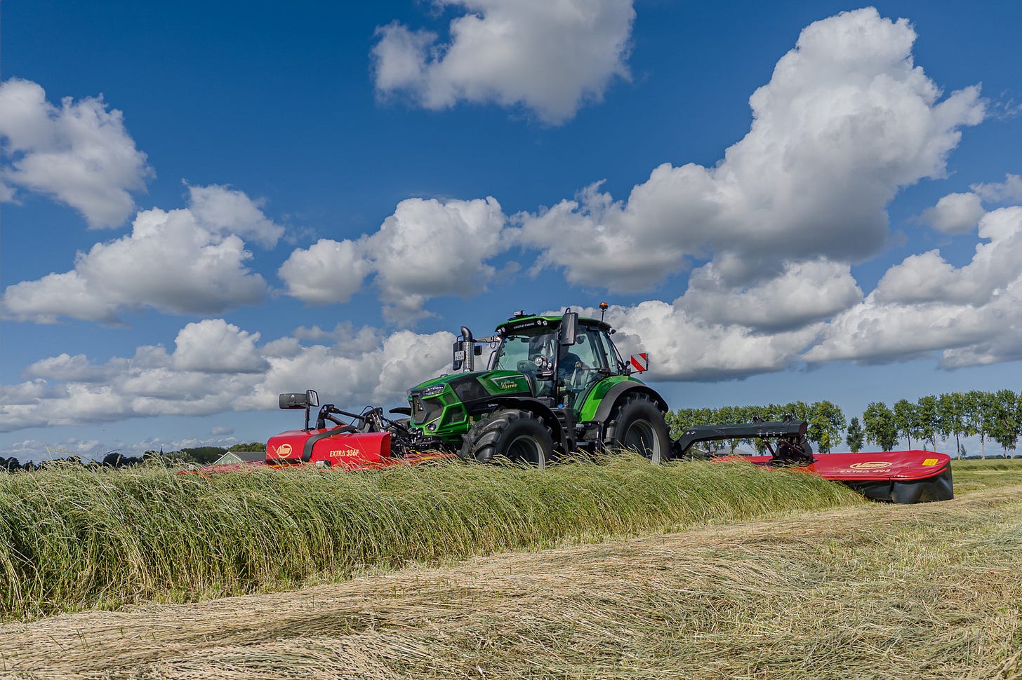 Ein grüner Deutz-Fahr-Traktor mit roter Mähkombination schneidet eine Wiese unter tiefblauem Himmel mit grossen Quellwolken. Vor dem Traktor steht noch hohes Gras, dahinter liegt das frisch gemähte Futter in breiten Schwaden auf dem Feld. Ein grüner Deutz-Fahr-Traktor mit roter Mähkombination schneidet eine Wiese unter tiefblauem Himmel mit grossen Quellwolken. Vor dem Traktor steht noch hohes Gras, dahinter liegt das frisch gemähte Futter in breiten Schwaden auf dem Feld.