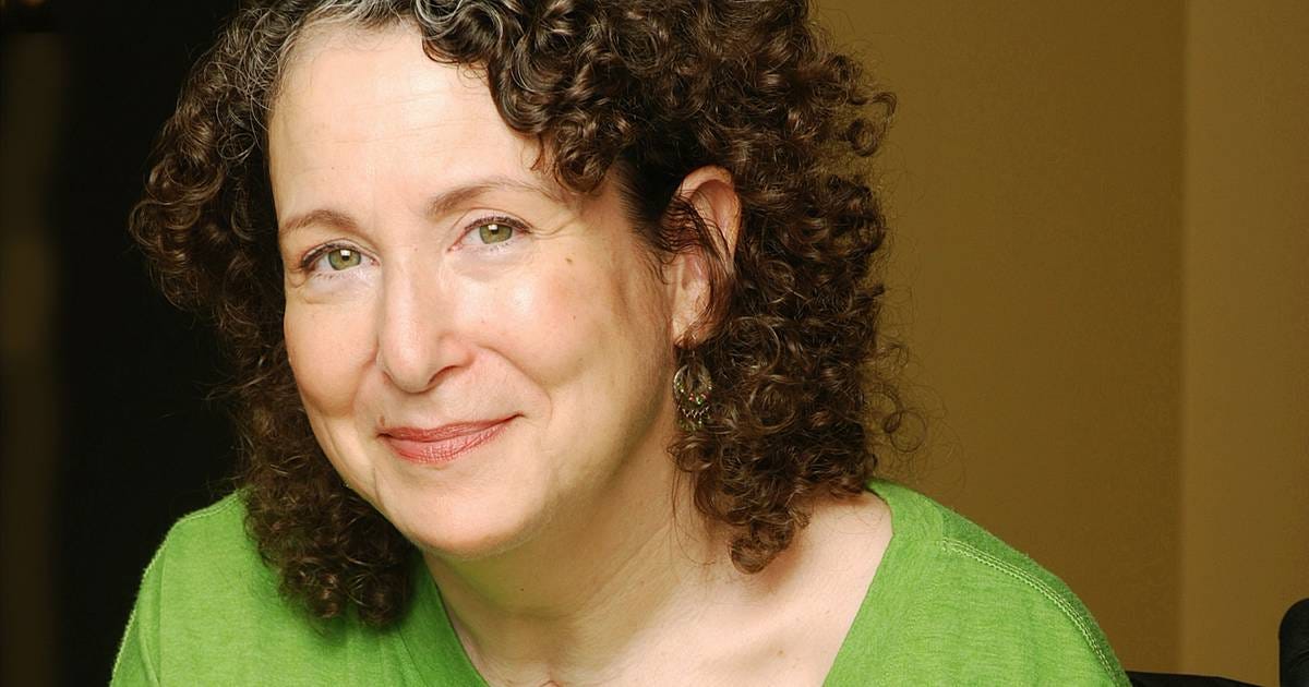 A white woman with curly brown hair and some grey at the roots is looking at the camera with a subtle, knowing smile.