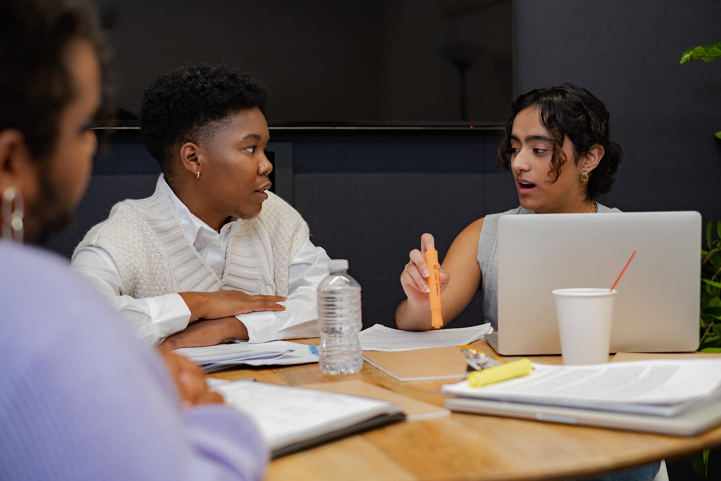 Three trans people around a table having a meeting; one has a laptop and an orange highlighter, one is crossing their arms behind a water bottle, and one is visible only from the side.