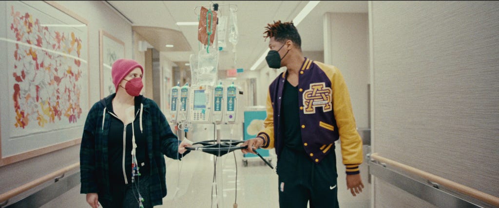 Suleika Jaouad and Jon Batiste in a hospital hallway, she in a pink hat and mask and hooked to a machine