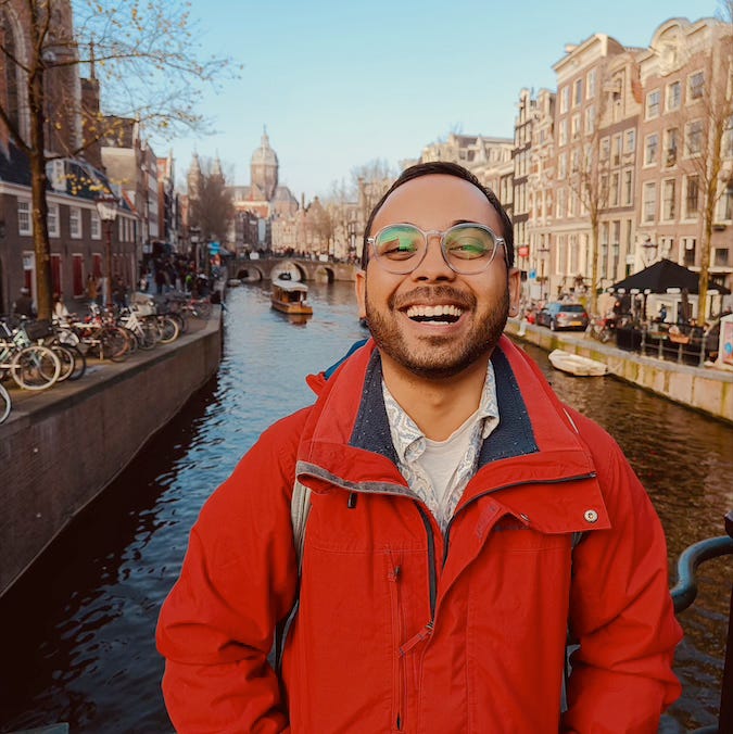 Siddharth, a young Indian man with black hair and stubble is dressed in a red jacket and layers of clothing within. He is wearing glasses with a round pale white frame and standing against a bridge on the Oudezijds Achterburgwal Canal in Amsterdam, laughing into the camera.
