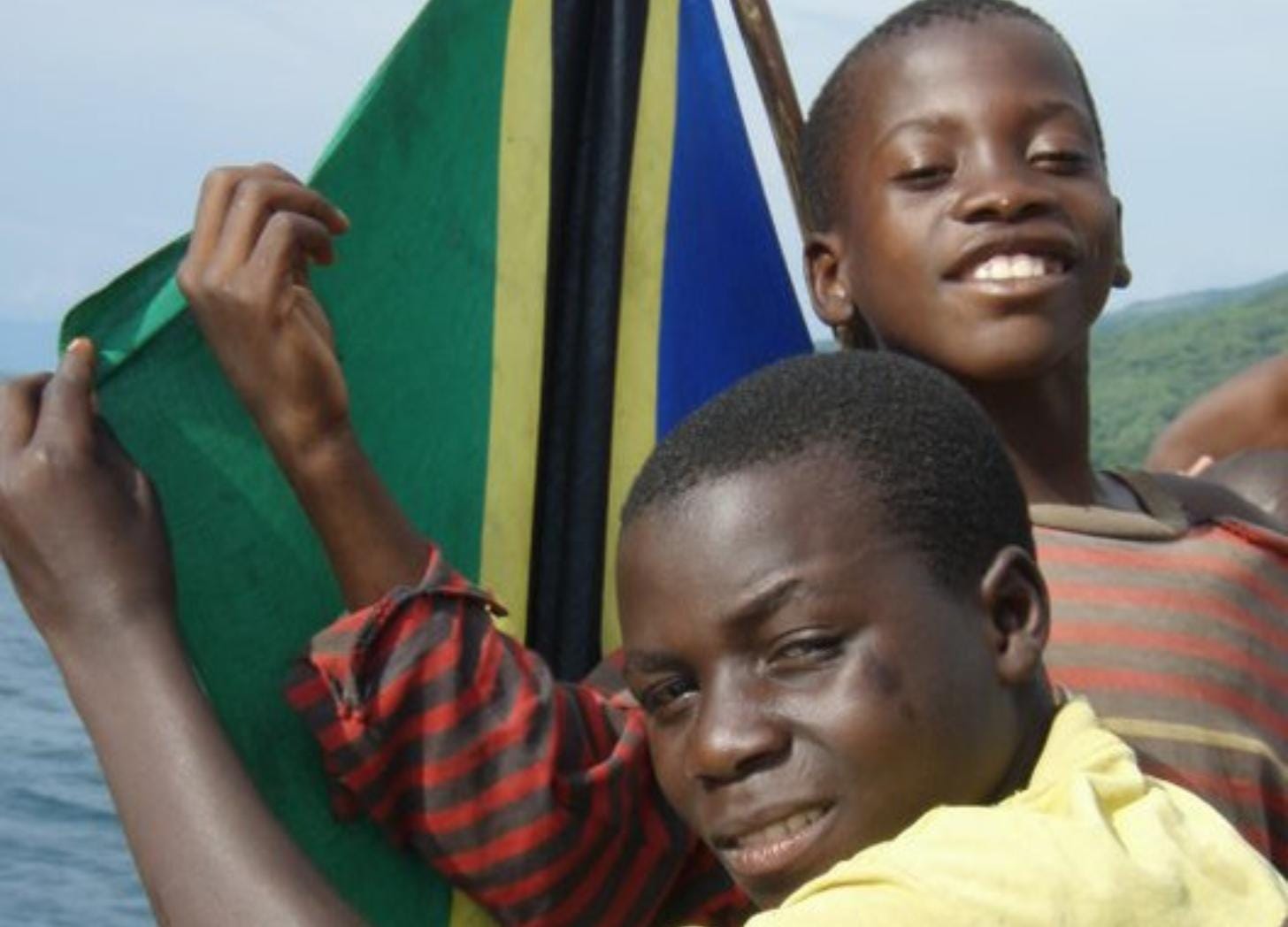 Two Tanzanian boys smile and hold up the Tanzania flag on a wooden boat rail, Lake Tanganyika behind them. Two Tanzanian boys smile and hold up the Tanzania flag on a wooden boat rail, Lake Tanganyika behind them.